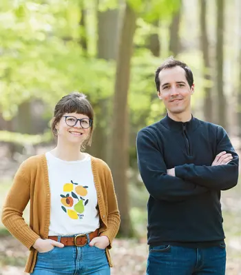 Zwei lächelnde Personen stehen im sonnigen Wald und schauen in die Kamera. Freundliche, authentische Porträtaufnahme in natürlicher Umgebung mit frischem Frühlingslicht.